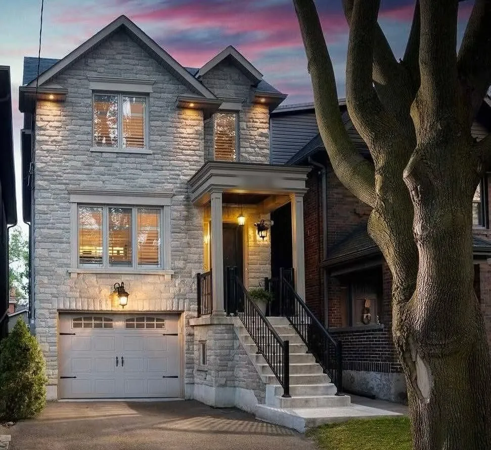 A narrow stone house at dusk with its porch and garage lights on.