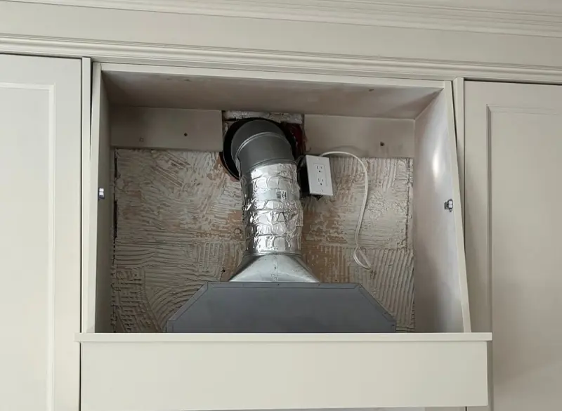 An open cabinet box over a stove with exposed duct work and no fan.